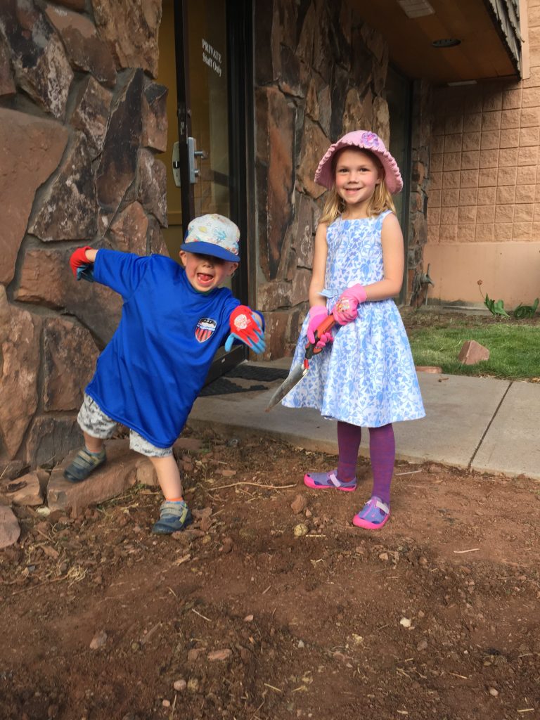 Two children smiling at the camera working in a garden.