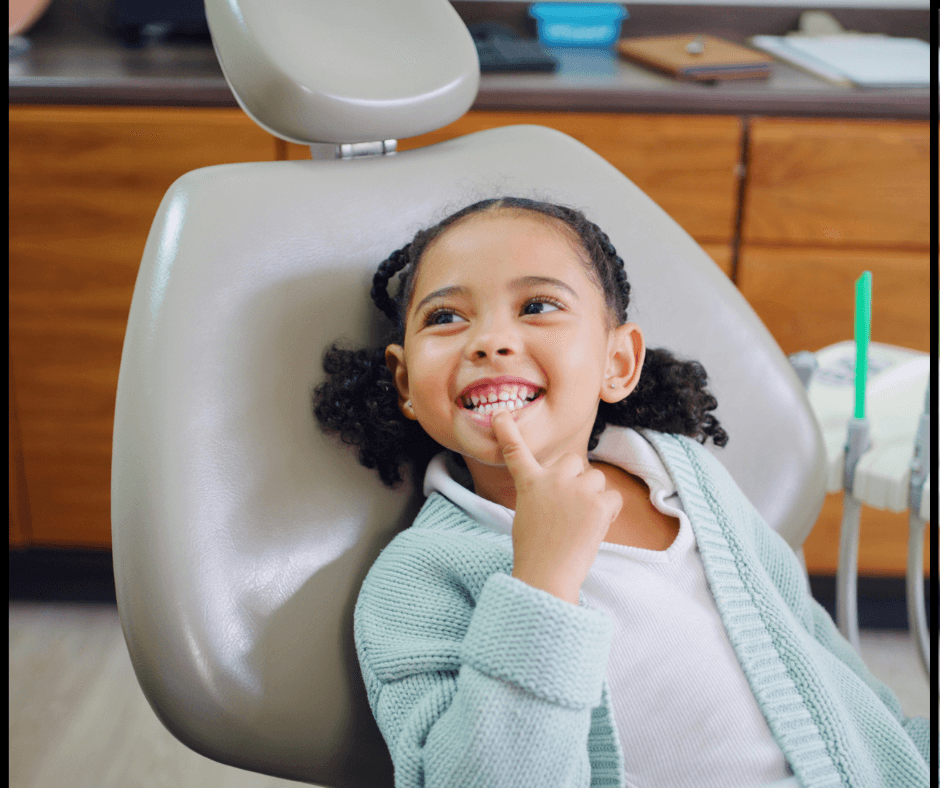 A child is sitting in a dental chair, smiling and pointing at their teeth.